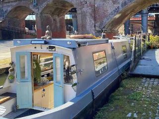 A houseboat moored under an arch of a brick bridge at Castlefield canals, adorned with plants and reflecting the city's canal-side living and industrial history
